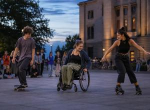 Riteņkrēsla lietotāja, skeiteris un skrituļotāja kopīgi kustas pilsētas laukumā vakara pasākumā.