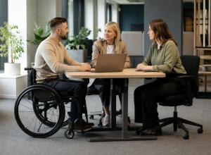 Riteņkrēsla lietotājs pie regulējama biroja galda pārrunā darba uzdevumus ar vadītāju un kolēģi.