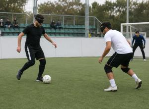 Divi spēlētāji ar acu aizsegiem sacenšas par bumbu neredzīgo futbola laukumā ar sānu apmalēm.