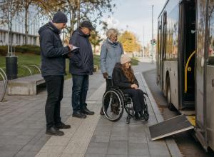 Riteņkrēsla lietotāja un trīs cilvēki pārbauda pieejamību pie pilsētas autobusa pieturas ar izlaistu rampu.