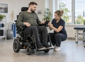 Elektriskā riteņkrēsla lietotājs kopā ar ergoterapeiti regulē vadības ierīci rehabilitācijas telpā.