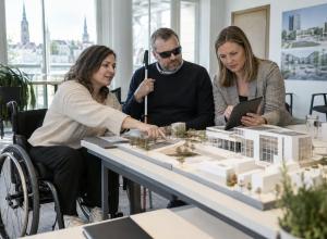 Riteņkrēsla lietotāja, cilvēks ar balto spieķi un speciāliste kopā izvērtē ēkas maketu un planšeti.