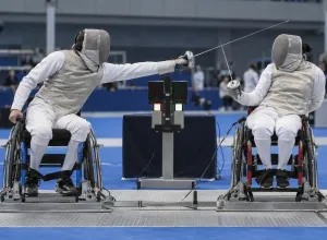 Divi para paukotāji nostiprinātos riteņkrēslos sacenšas uz metāla rāmja sporta hallē
