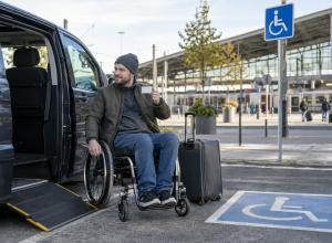 Ceļotājs riteņkrēslā pie pielāgota auto un dzelzceļa stacijas tur rokā karti