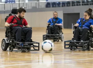 Trīs sportisti elektriskajos riteņkrēslos kontrolē lielu futbola bumbu sporta zālē