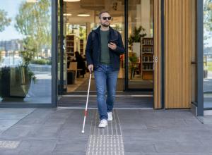 Neredzīgs cilvēks ar balto spieķi un tālruni pārvietojas pa taktilo vadlīniju pie bibliotēkas ieejas