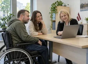 Cilvēks riteņkrēslā ar tuvinieci un sociālo darbinieci pie galda pārskata informāciju datorā