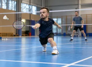 Maza auguma sportists izklupienā atsit volānu badmintona laukumā, treniņu partnerim stāvot fonā