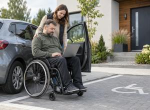Cilvēks riteņkrēslā pie automašīnas lieto datoru kopā ar atbalsta personu