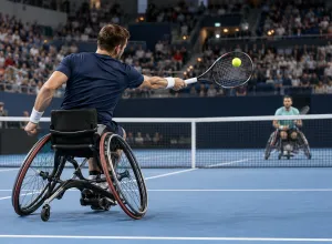 Divi riteņtenisisti sporta riteņkrēslos sacenšas uz zilā seguma laukuma pilnā iekštelpu arēnā