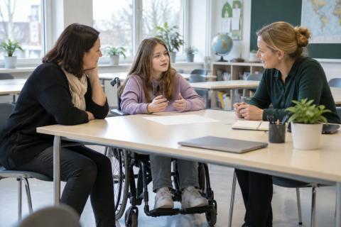 Skolniece riteņkrēslā kopā ar vecāku un skolotāju pārrunā mācību plānu klasē