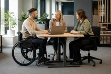Riteņkrēsla lietotājs pie regulējama biroja galda pārrunā darba uzdevumus ar vadītāju un kolēģi.