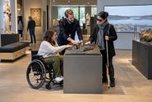 Riteņkrēsla lietotāja un apmeklētāja ar balto spieķi kopā ar muzeja darbinieku izpēta taustes maketu.