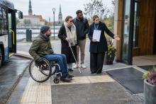 Riteņkrēsla lietotājs un cilvēks ar balto spieķi kopā ar tūrisma darbinieci pārbauda pieejamu ieeju.