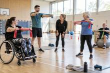 Dažādu paaudžu cilvēki sporta zālē kopīgi vingro ar elastīgajām lentēm