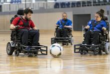Trīs sportisti elektriskajos riteņkrēslos kontrolē lielu futbola bumbu sporta zālē