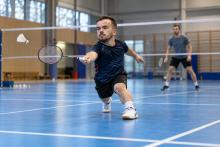 Maza auguma sportists izklupienā atsit volānu badmintona laukumā, treniņu partnerim stāvot fonā