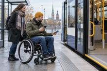 Vīrietis riteņkrēslā ar transporta karti un tālruni gatavojas iekāpt zemās grīdas sabiedriskajā transportā.