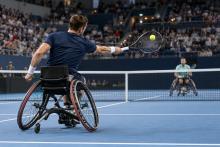 Divi riteņtenisisti sporta riteņkrēslos sacenšas uz zilā seguma laukuma pilnā iekštelpu arēnā
