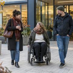 Sieviete riteņkrēslā sarunājas ar draugiem pie pieejamas kopienas ēkas