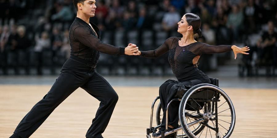 Para deju pāris sporta zālē izpilda sinhronu pagriezienu, vienam dejotājam izmantojot riteņkrēslu.
