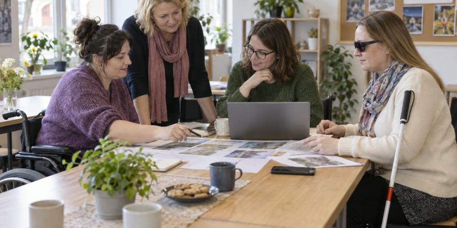 Sievietes ar invaliditāti kopīgi apspriež projekta plānu pie galda ar datoru un materiāliem