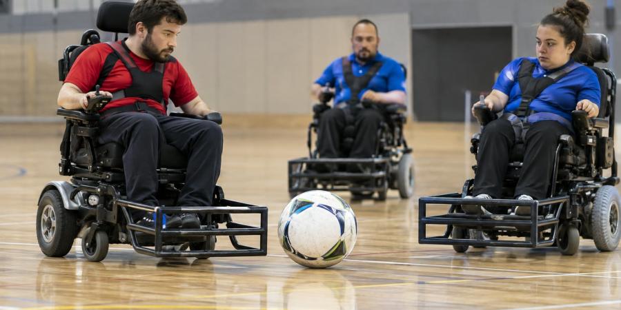 Trīs sportisti elektriskajos riteņkrēslos kontrolē lielu futbola bumbu sporta zālē
