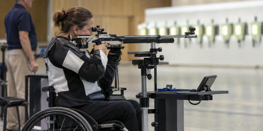 Sportiste riteņkrēslā mērķē ar atbalsta statīvā nostiprinātu šauteni iekštelpu šautuvē