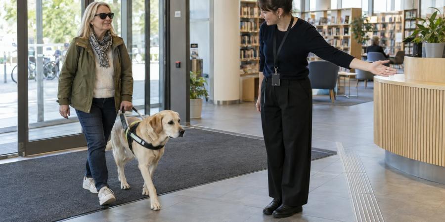 Sieviete ar asistējošo suni ieiet bibliotēkā, kur darbiniece norāda brīvu ceļu