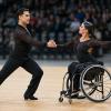 Para deju pāris sporta zālē izpilda sinhronu pagriezienu, vienam dejotājam izmantojot riteņkrēslu.