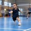 Maza auguma sportists izklupienā atsit volānu badmintona laukumā, treniņu partnerim stāvot fonā