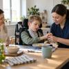 Aprūpes speciāliste palīdz bērnam pie galda, kamēr mamma vēro viņu ikdienas gaitas
