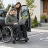Cilvēks riteņkrēslā pie automašīnas lieto datoru kopā ar atbalsta personu
