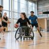 Parasportisti kopā ar treneri izpilda bumbas vingrinājumu gaišā un pieejamā sporta zālē.