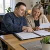 Cilvēks riteņkrēslā kopā ar tuvinieci pie galda pārskata aploksni un sakārtotu dokumentu mapi