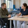 Cilvēks riteņkrēslā rehabilitācijas kabinetā pārrunā individuālo plānu ar speciālisti