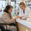 Sieviete riteņkrēslā kopā ar farmaceiti pie letes pārskata zāļu iepakojumu un lietošanas plānu