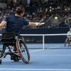 Divi riteņtenisisti sporta riteņkrēslos sacenšas uz zilā seguma laukuma pilnā iekštelpu arēnā