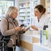 Aptiekā farmaceite izskaidro zāļu iepakojumu klientei, kura izmanto staigāšanas rāmi