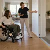Sieviete riteņkrēslā kopā ar speciālisti vērtē plašu ieeju un atbalsta rokturi mājoklī